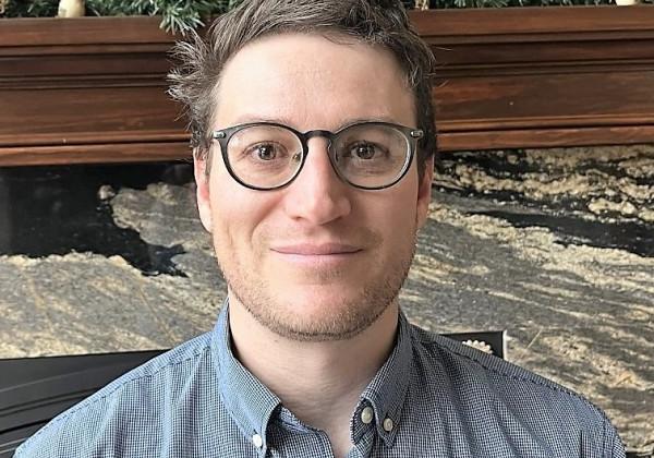 A man with brunette hair with glasses smiling at camera. 