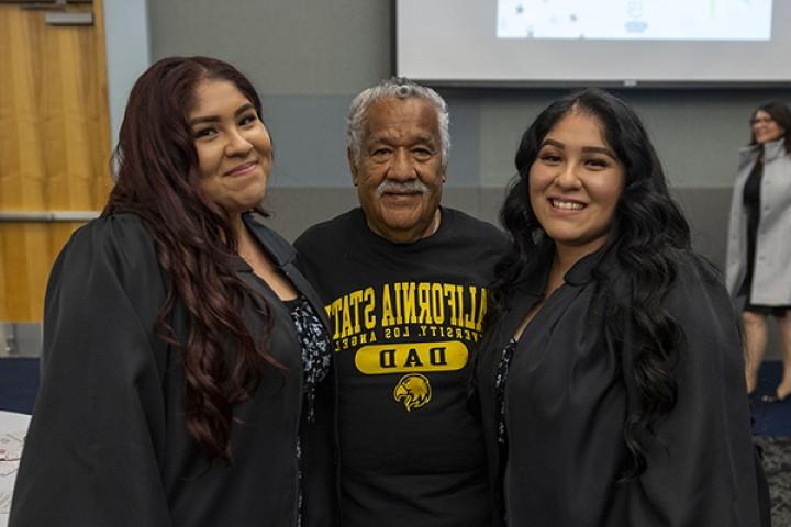 One person standing in between two students who are wearing graduation gowns.