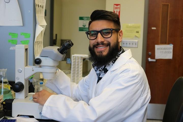 male student with a beard, glasses and white lab coat smiling at the camera. 他在实验室里拿着显微镜