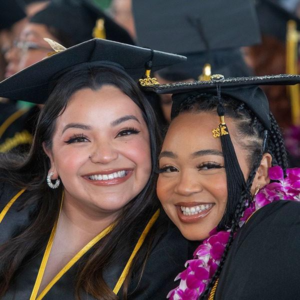 2 graduates at commencement, sitting cheek-to-cheek