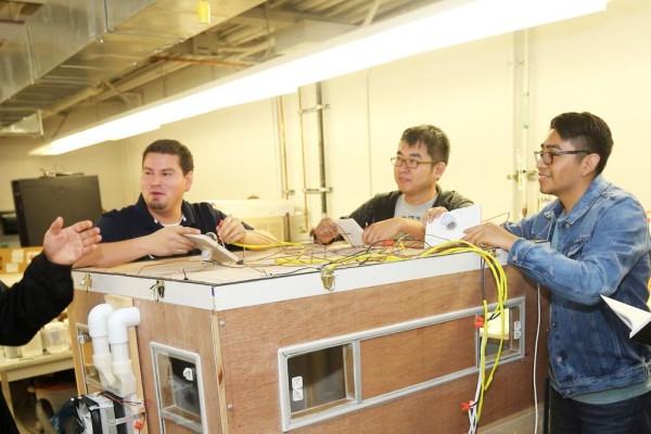 Image of a group of engineering students surrounding a large wooden box device while their professor addresses them. 