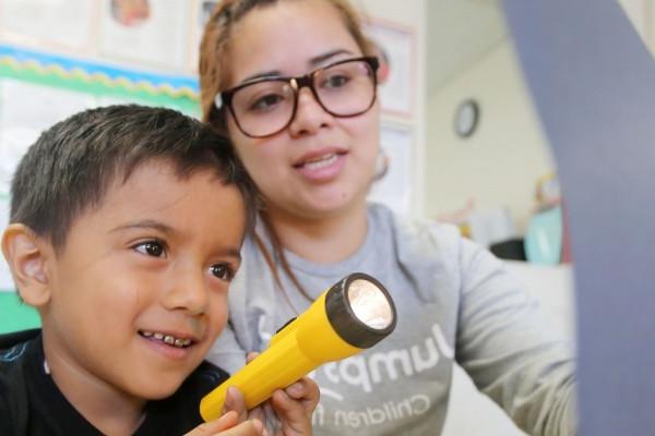 Image of a female student holding up a page for a child who is shining a flashlight on it. 