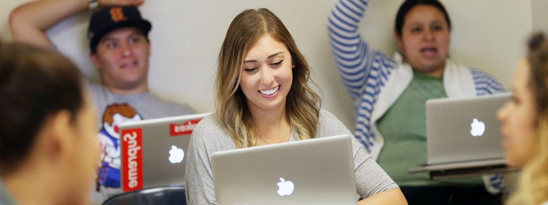 Image of a Caucasian female student smiling in a classroom setting in front of an open laptop. 