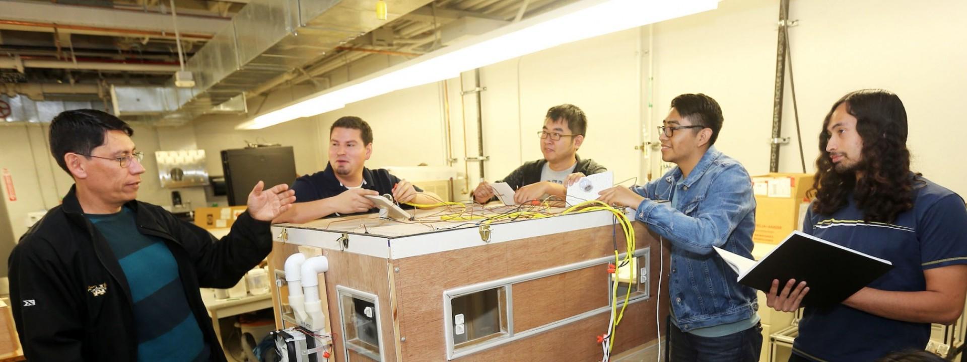 Image of a group of engineering students surrounding a large wooden box device while their professor addresses them. 