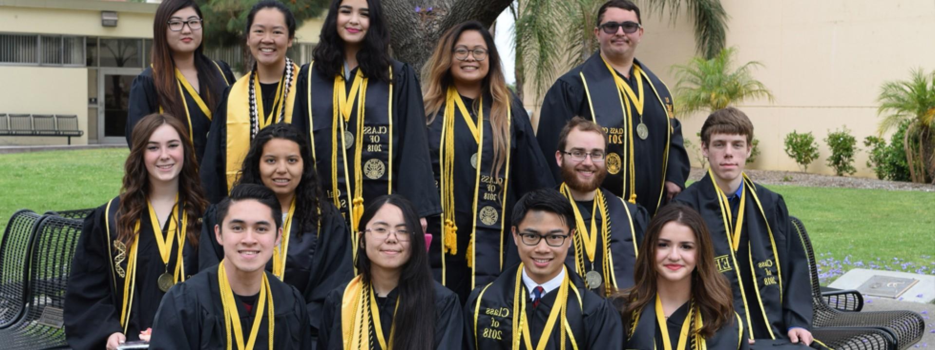 Group of Honors College graduating students in Commencement regalia, gathered in front of the jacaranda tree on campus. 